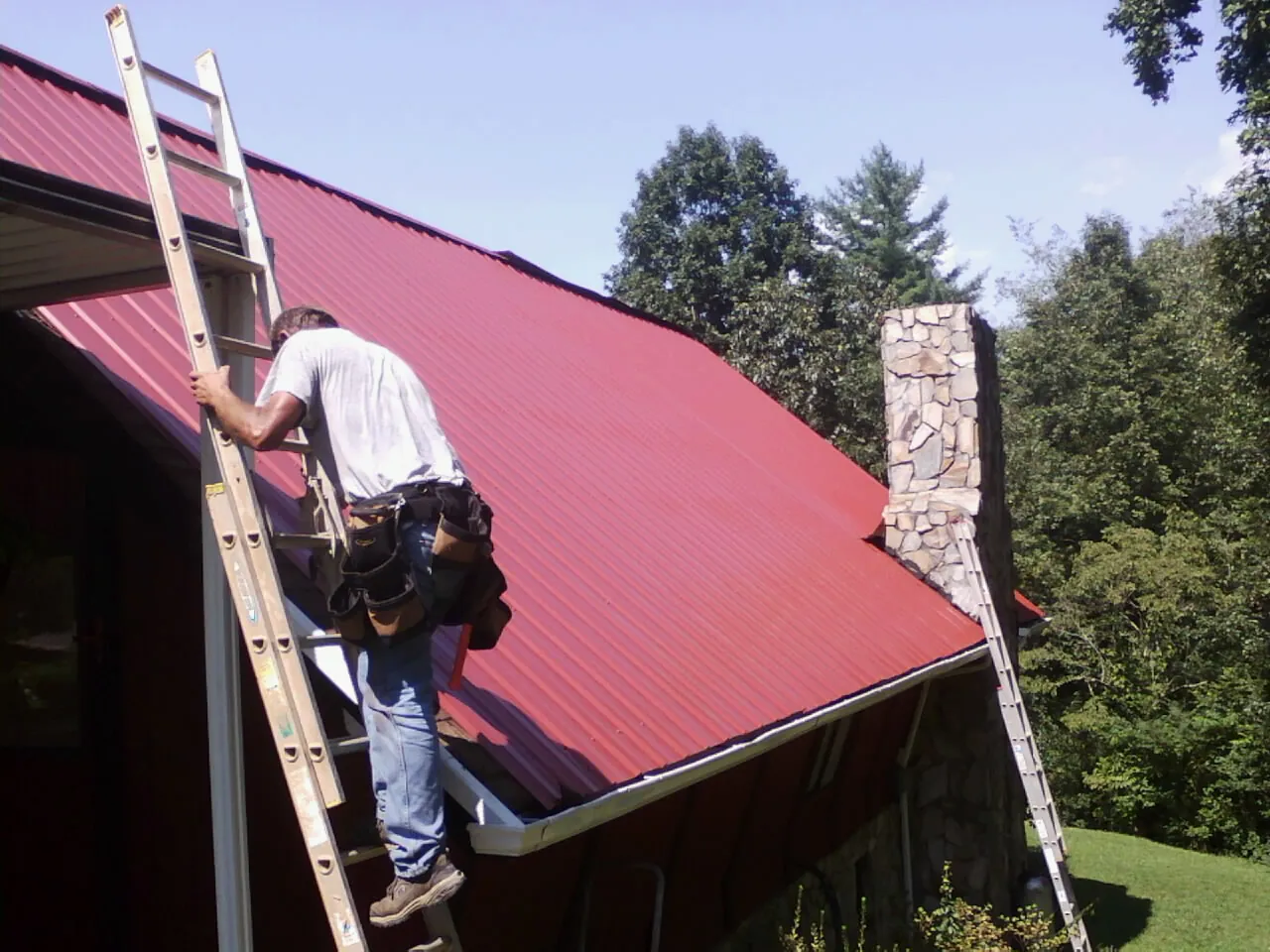 Roofer on a ladder beside a red metal roof and stone chimney — Soffit & Fascia Repair by Roofing Brothers in Brownsville